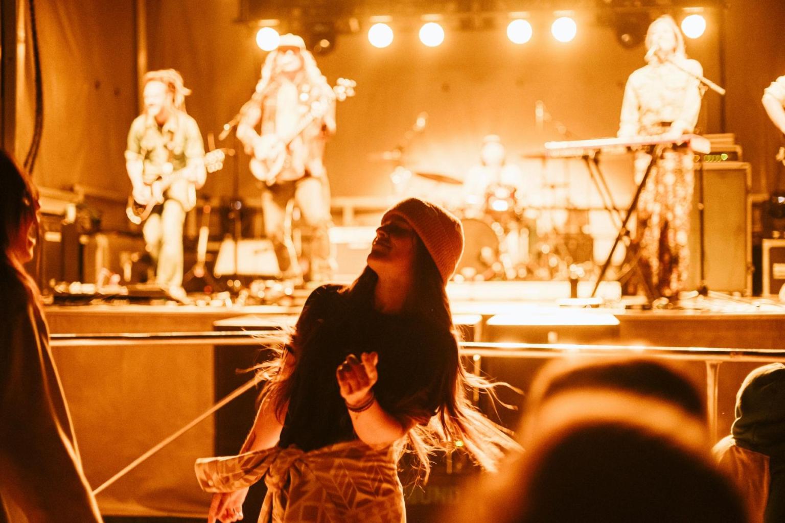 Concert with a band onstage and a person dancing in front, backlit by warm lights.