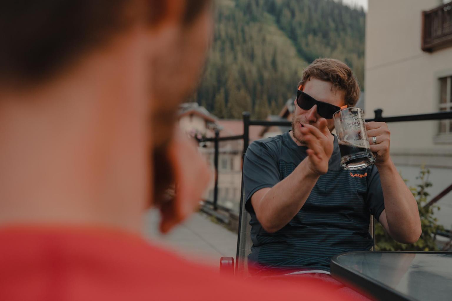 Man in sunglasses holds beer mug, gesturing on a balcony.