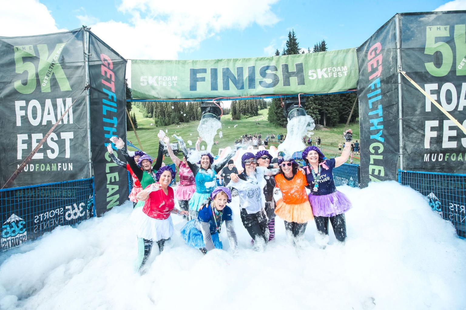 Participants celebrate in foam at a 5K race finish line, smiling and waving.