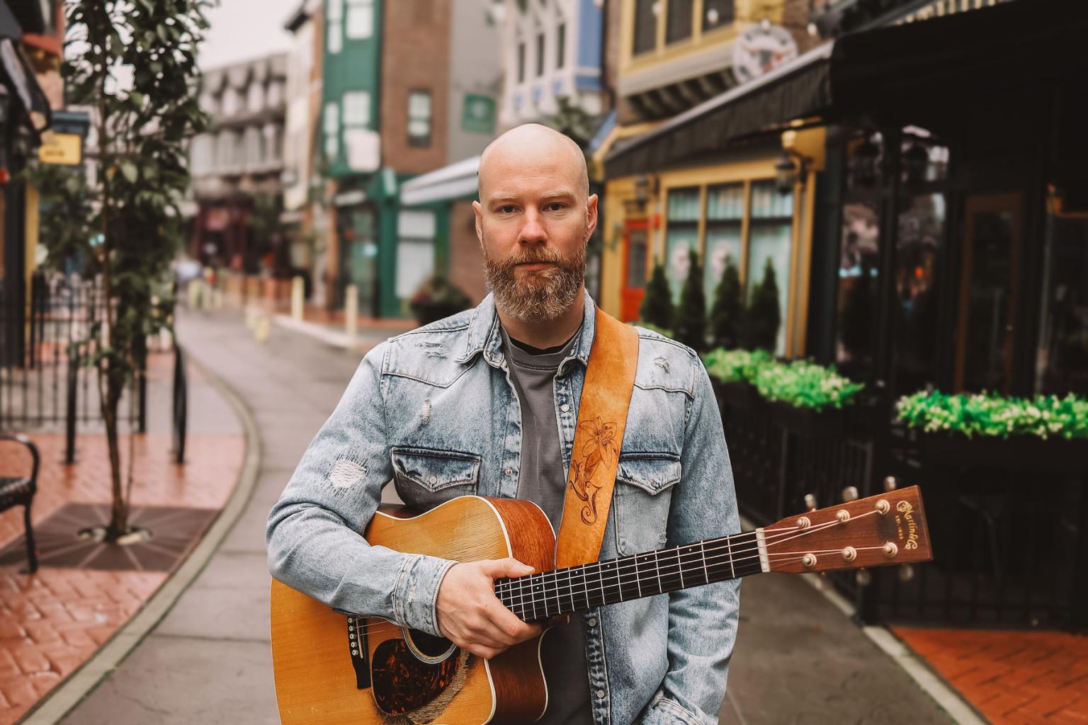 A man in a denim jacket holds a guitar on a cobblestone street.