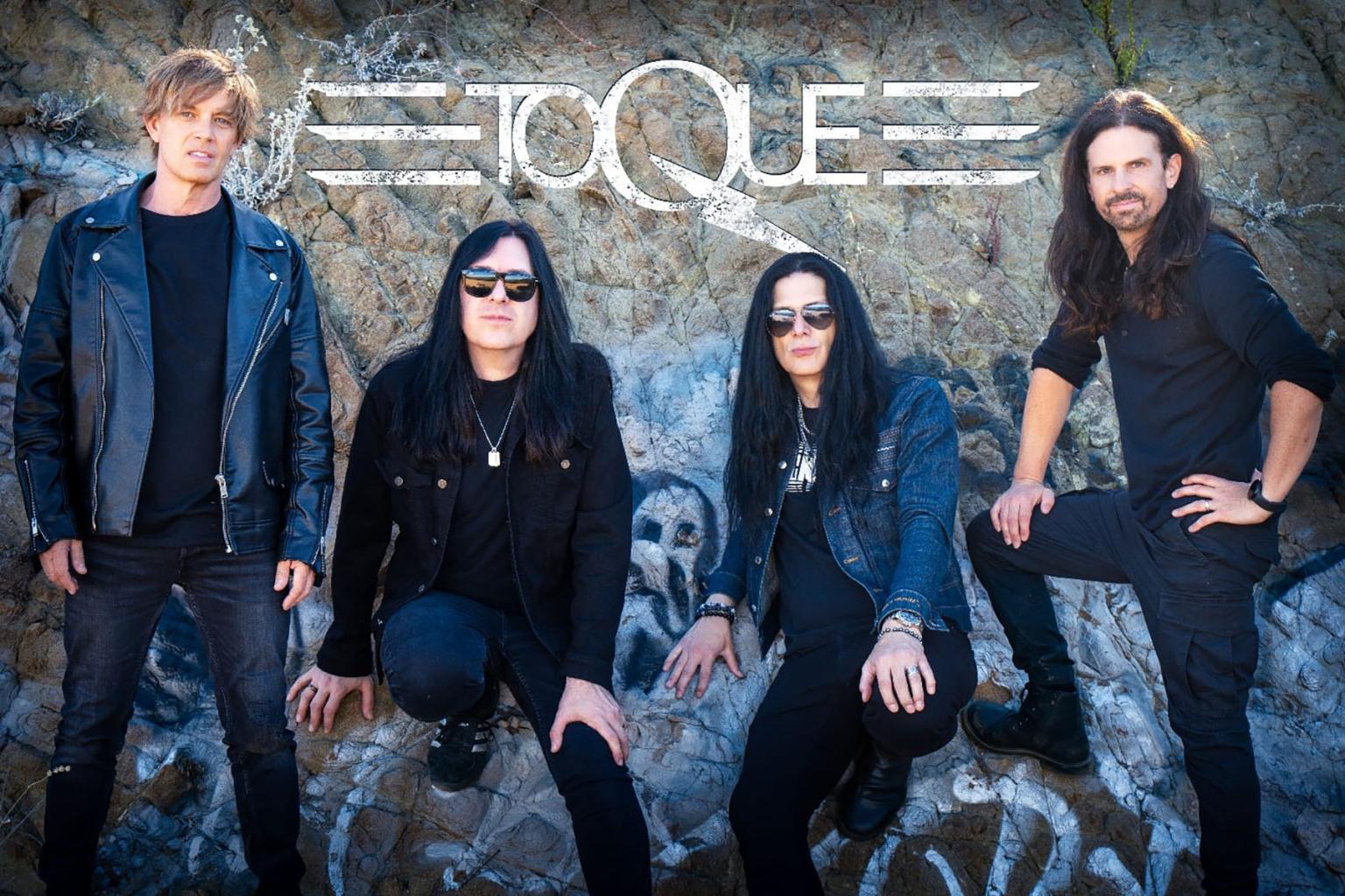 Four men with long hair stand and sit in front of a graffiti wall.