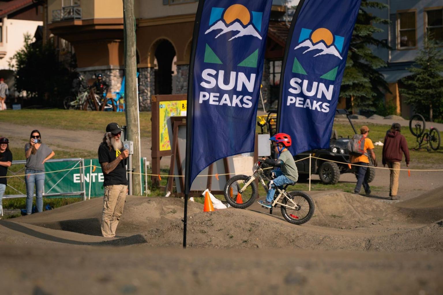 Child on a small dirt bike track under flags at a Sun Peaks event.