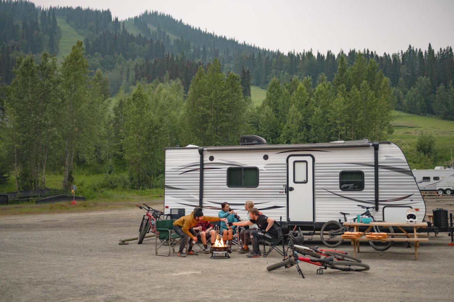 Camper with people sitting by a campfire, surrounded by trees and mountains.