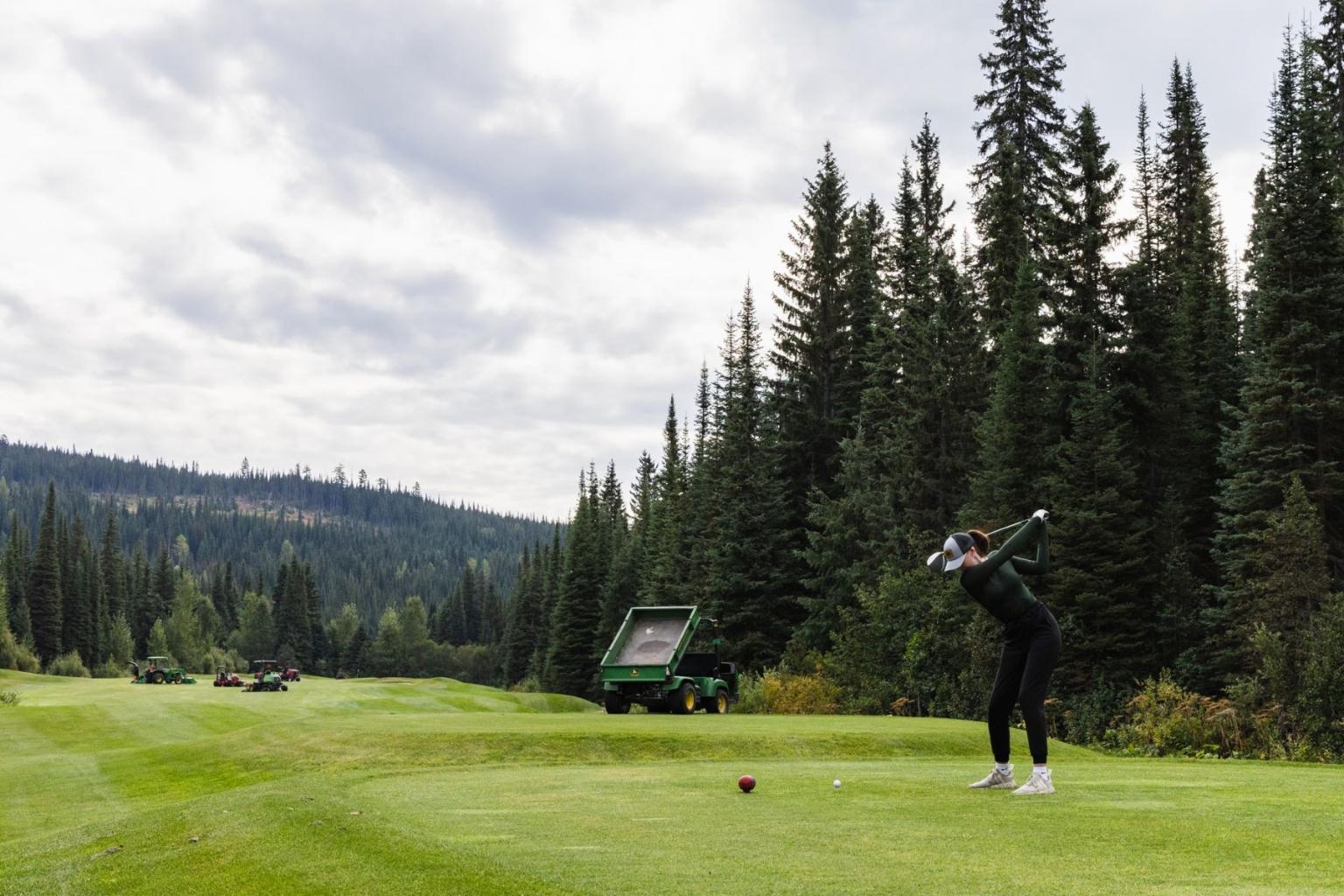 Golfer swinging on lush course with machinery in the middle and tall evergreens and cloudy sky.