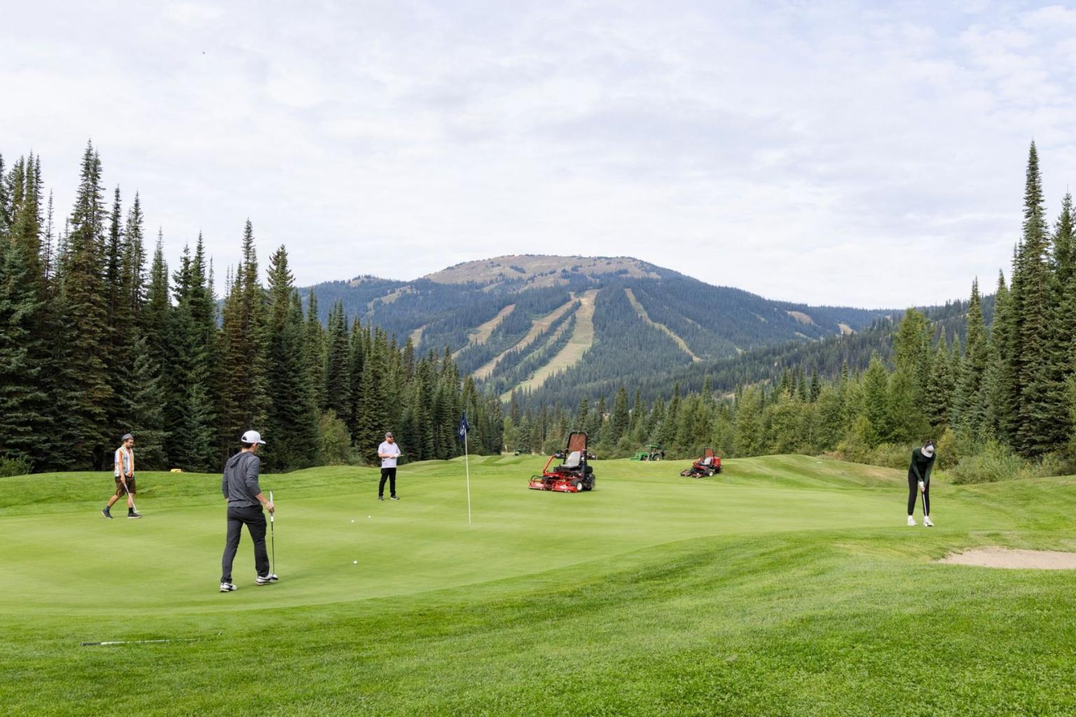 Golfers on a green course with machinery in the middle, and with mountains and trees in the background.