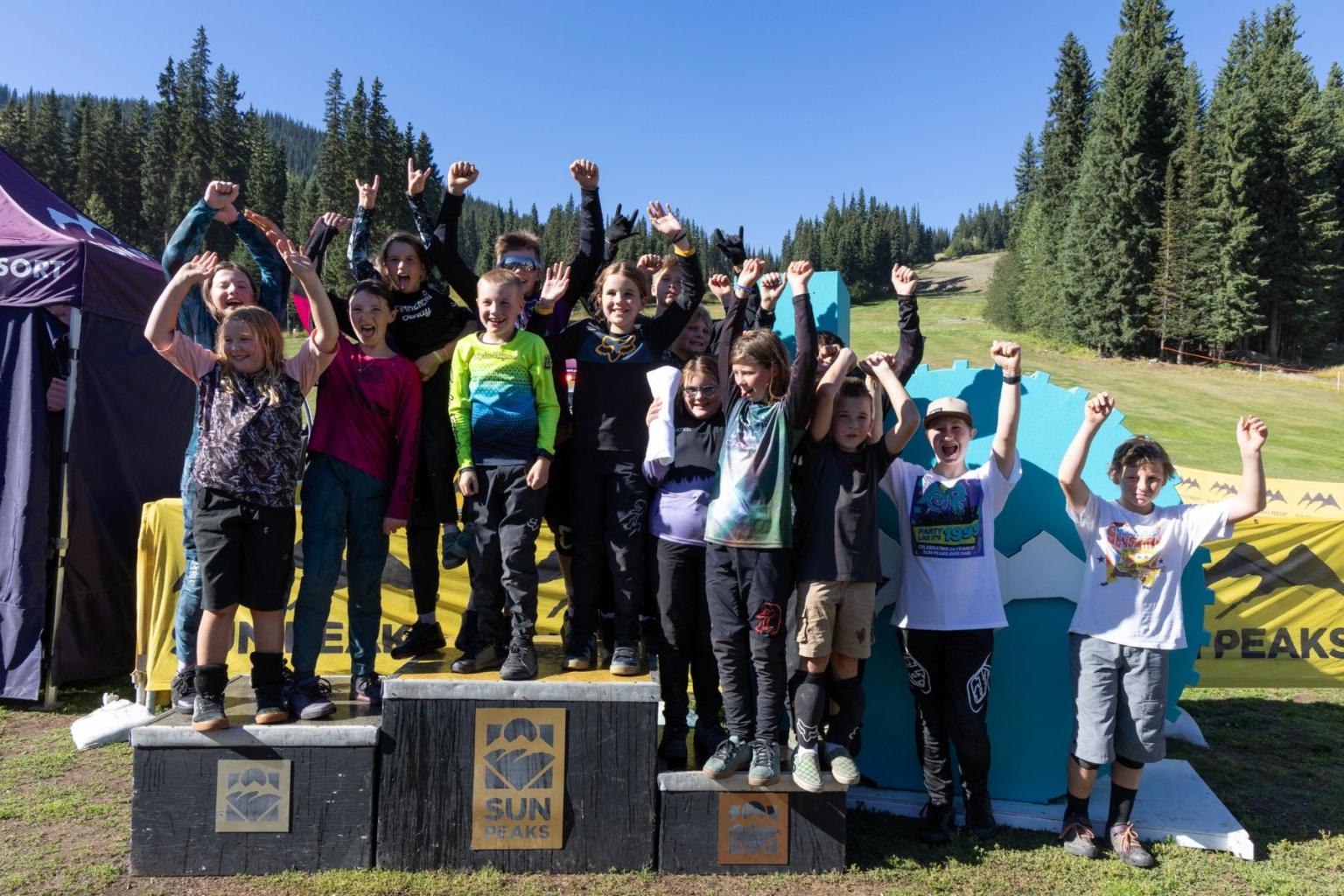 Children standing on a podium, cheering in a sunny outdoor setting with trees and tents after the Junior DH event.
