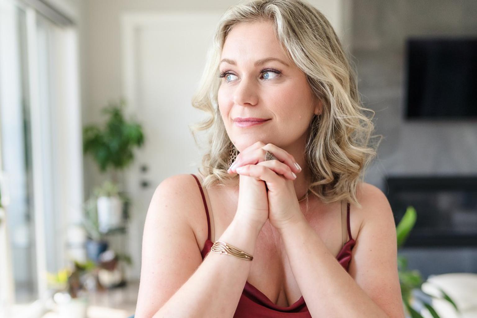 Smiling woman with clasped hands in a bright room, wearing a sleeveless top.