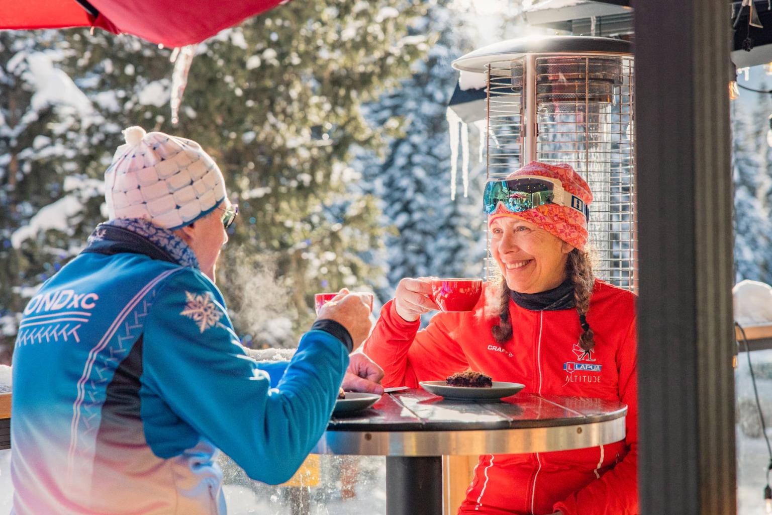 Two people in winter gear enjoy hot drinks outside on a sunny, snowy day.