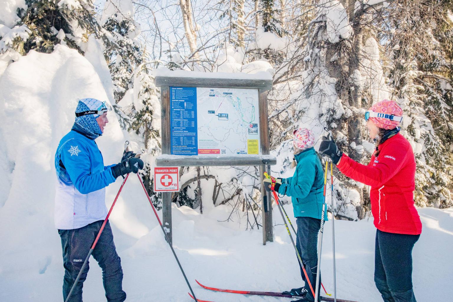 Three people in winter gear read a snowy trail map, surrounded by trees.
