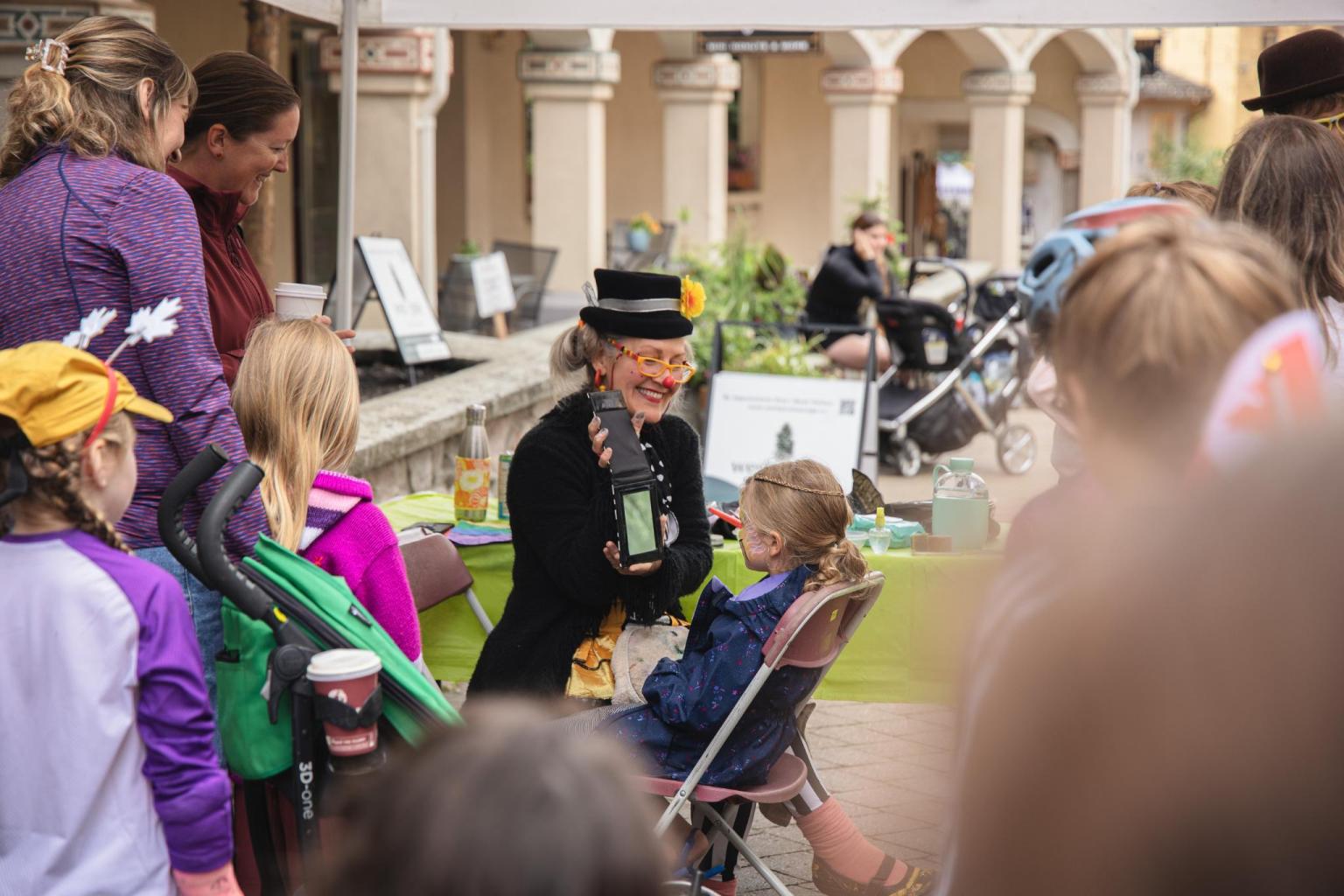 A clown entertains a seated child outdoors, surrounded by onlookers.