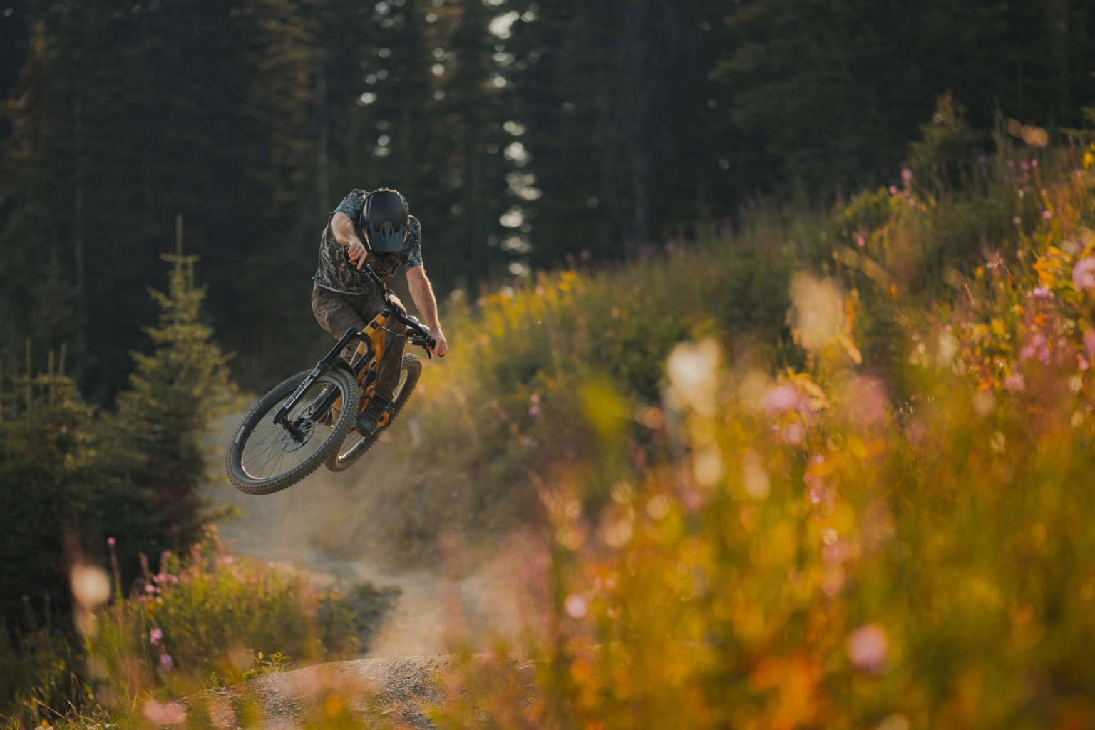 Mountain biker jumping on a dirt trail amidst wildflowers and trees.