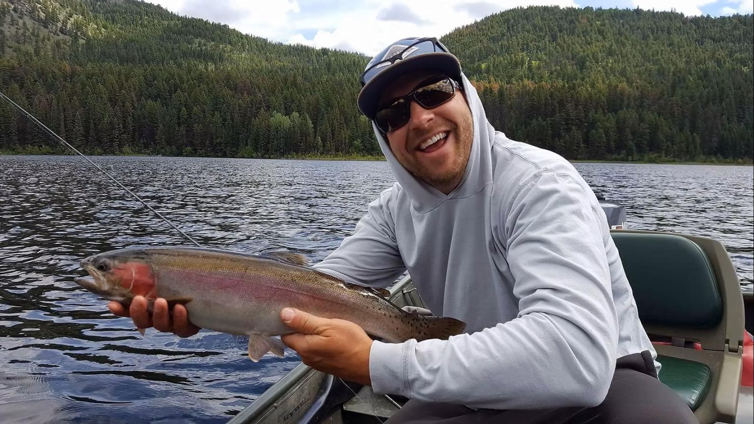 Man smiling while holding a large fish on a boat, with a forested lake backdrop.