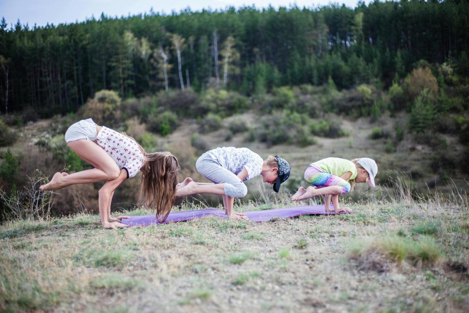 Children practicing yoga on mats in a grassy field, trees in the background.