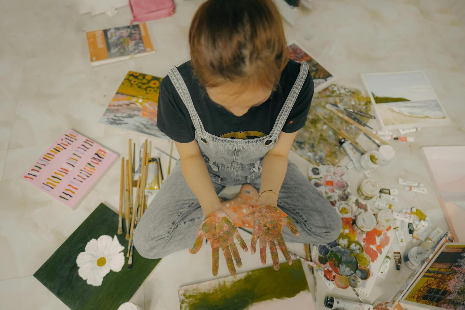 Child with paint-covered hands sits on floor surrounded by art supplies and paintings.