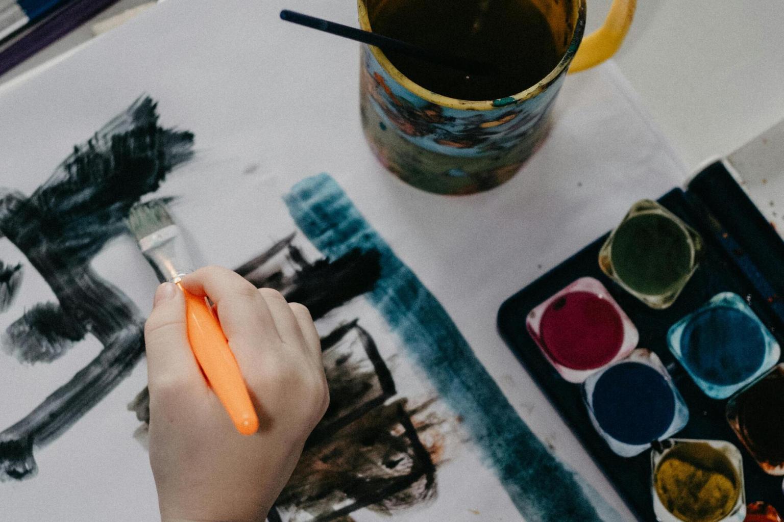 Hand painting with watercolors, paint jars nearby.