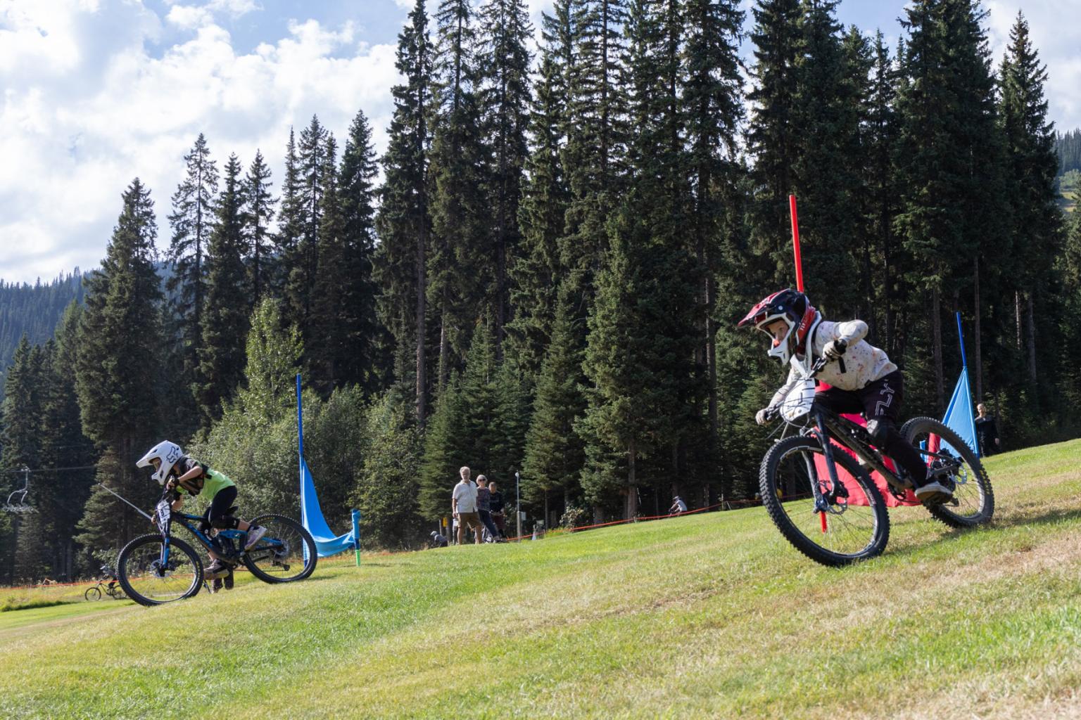 Two cyclists racing downhill on a grassy slope with tall trees in the background.