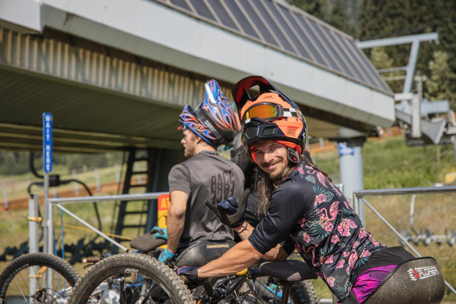 Two people in biking gear and helmets, smiling near a building.