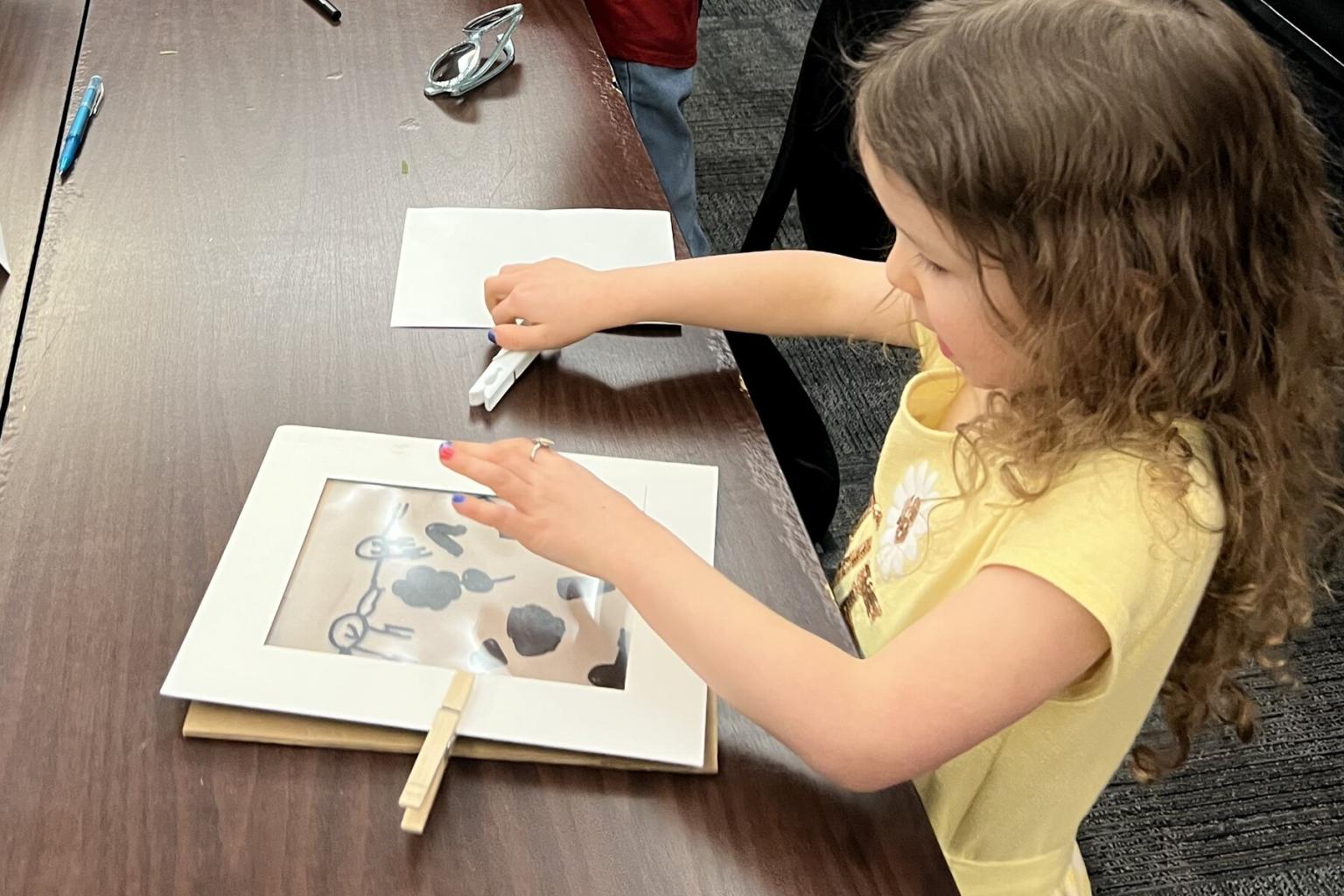 Young girl drawing on a paper with shapes, seated at a table.