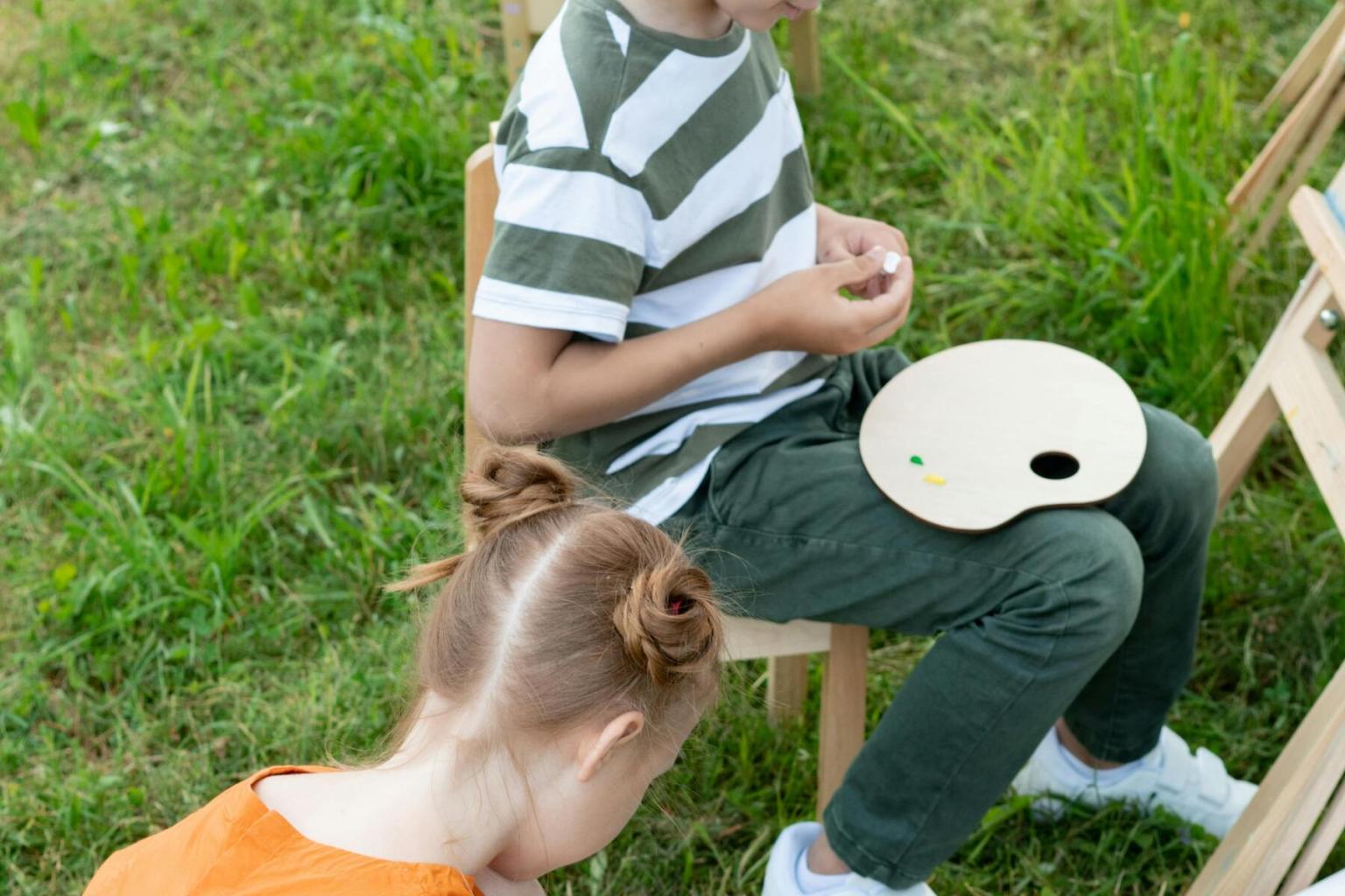 Child sitting on grass with a paint palette, another child nearby focused on work.