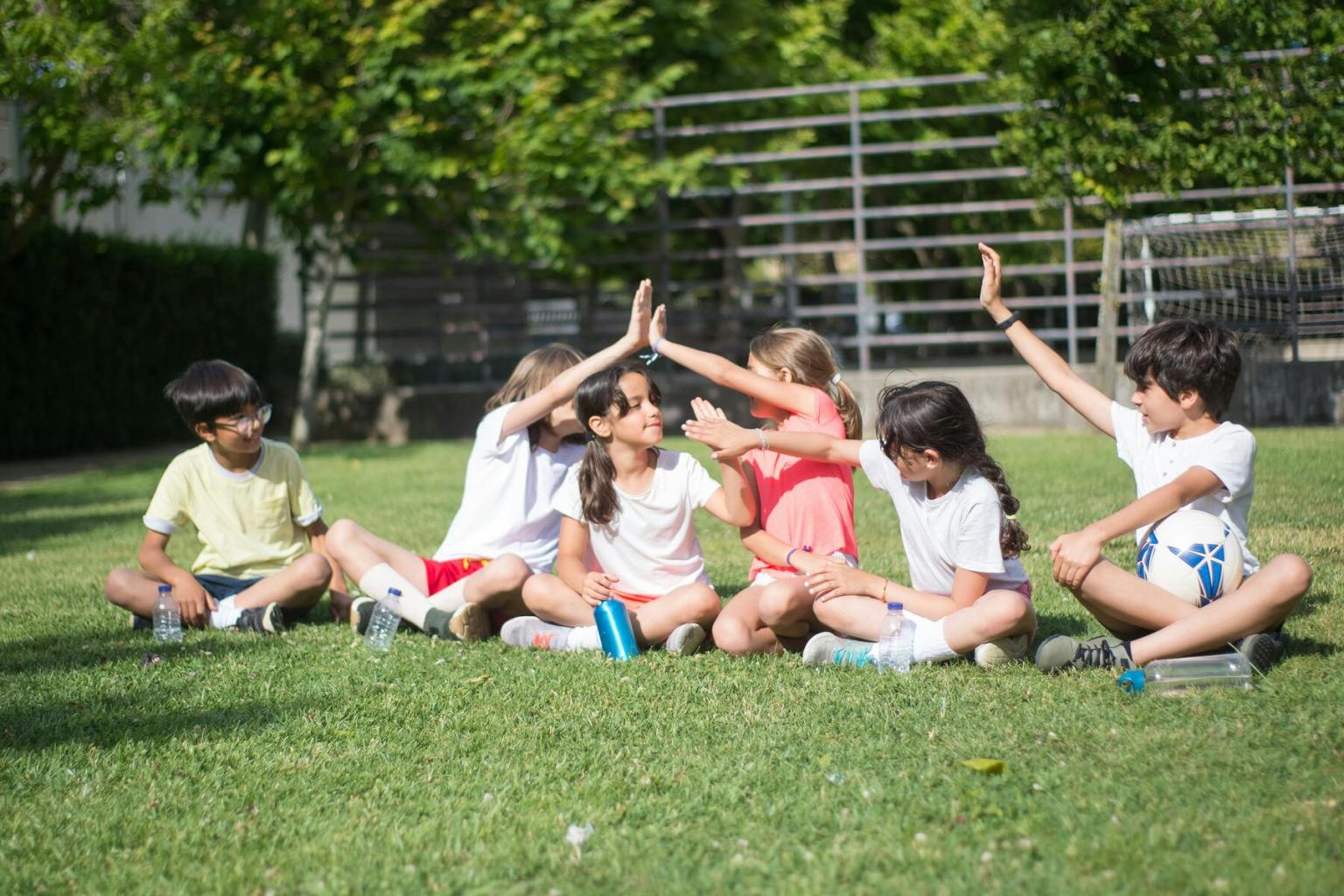 Kids sitting on grass, high-fiving in a sunny park.