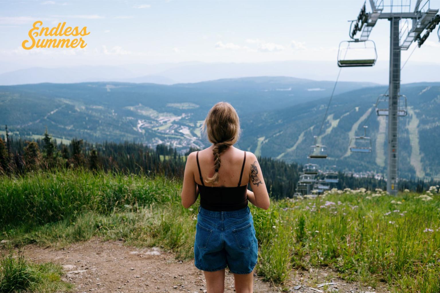 Woman in summer outfit overlooks mountains and chairlift on a clear day. Yellow Endless Summer cursive text.