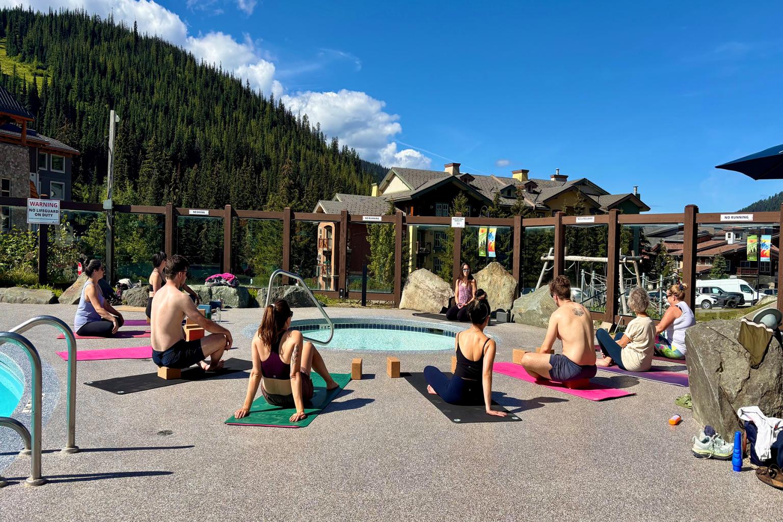 Outdoor yoga class by a pool, beneath a clear blue sky and mountains.