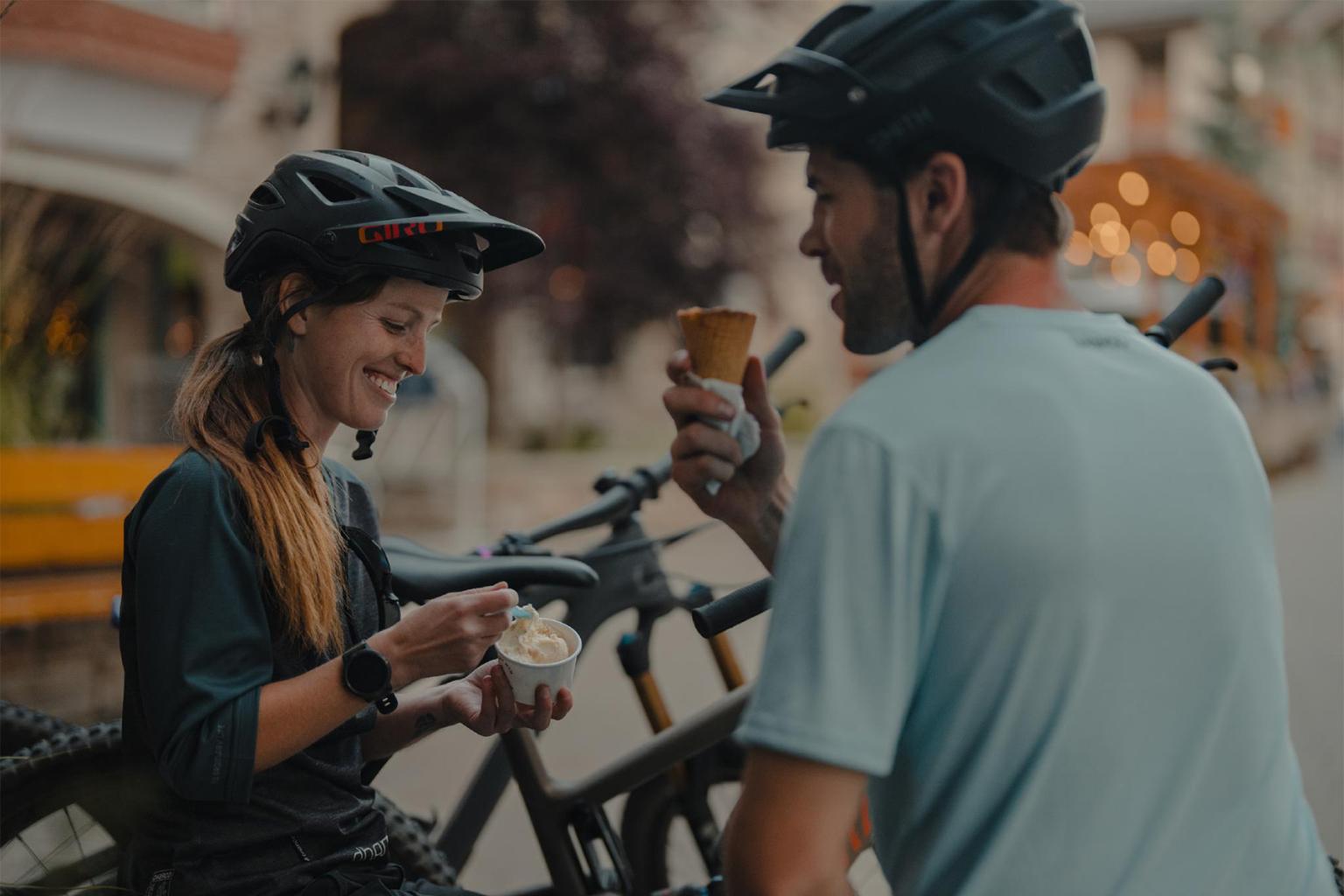 Two cyclists in helmets enjoy ice cream outdoors.
