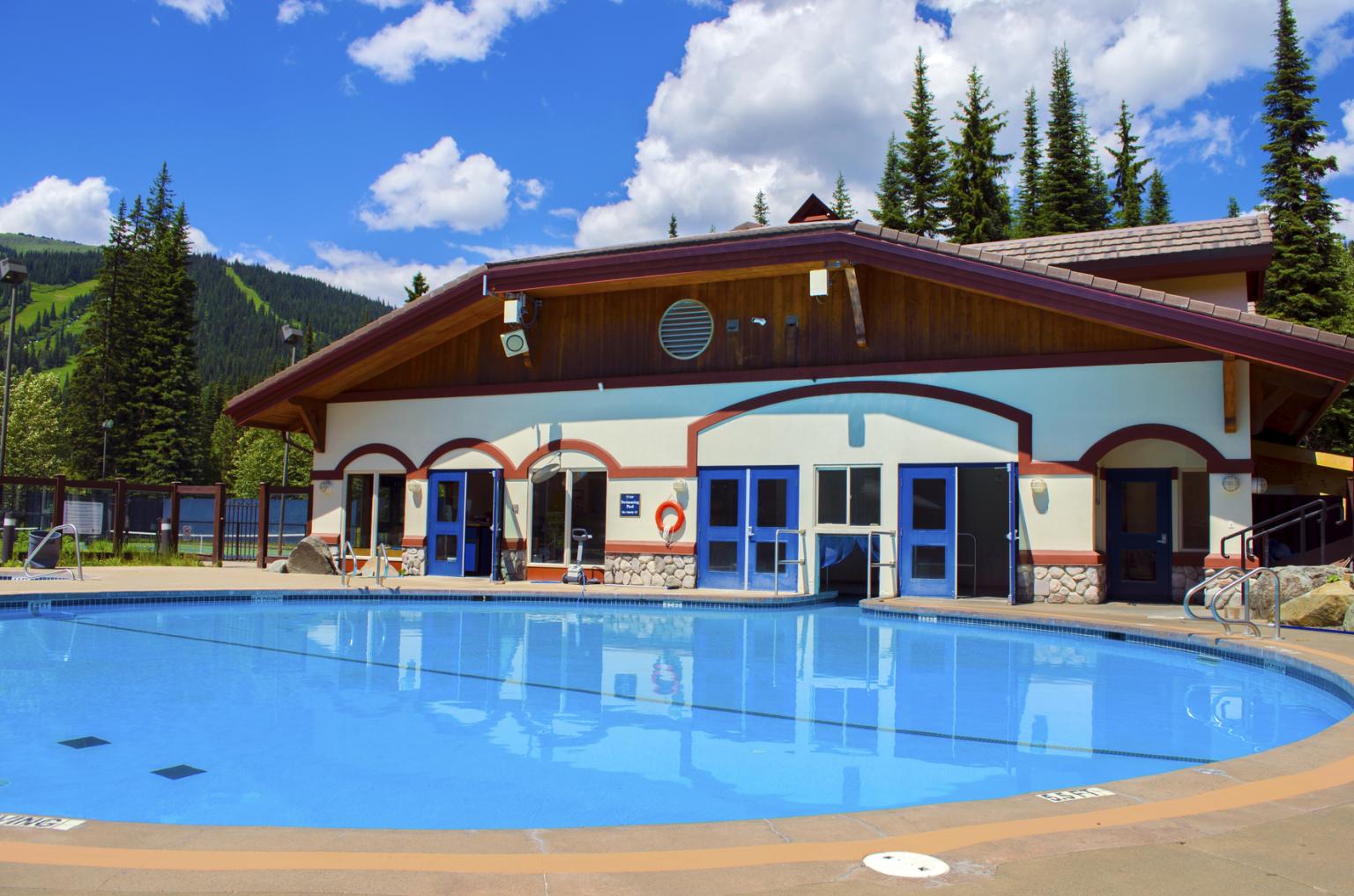 Outdoor pool with a wooden lodge, surrounded by trees and mountains under blue sky.