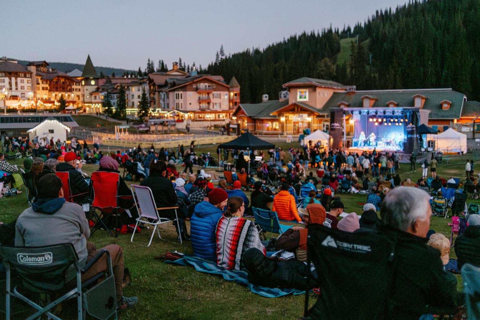 Outdoor concert at dusk with crowd on lawn chairs facing a lit stage.
