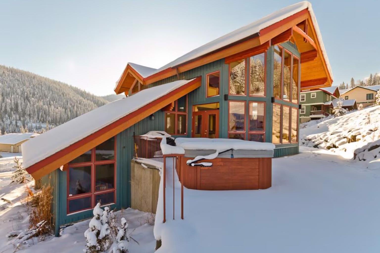 Snow-covered modern mountain cabin with large windows and a hot tub.