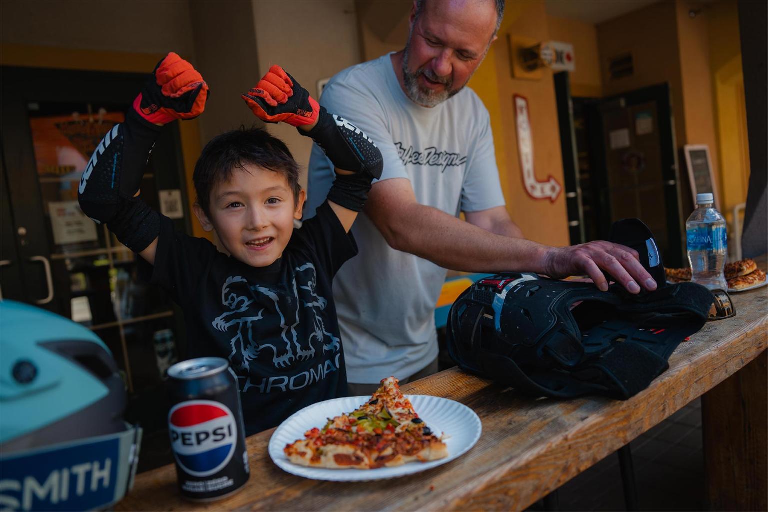 Child in protective gear celebrates with a man, pizza, and a soda on a wooden table.
