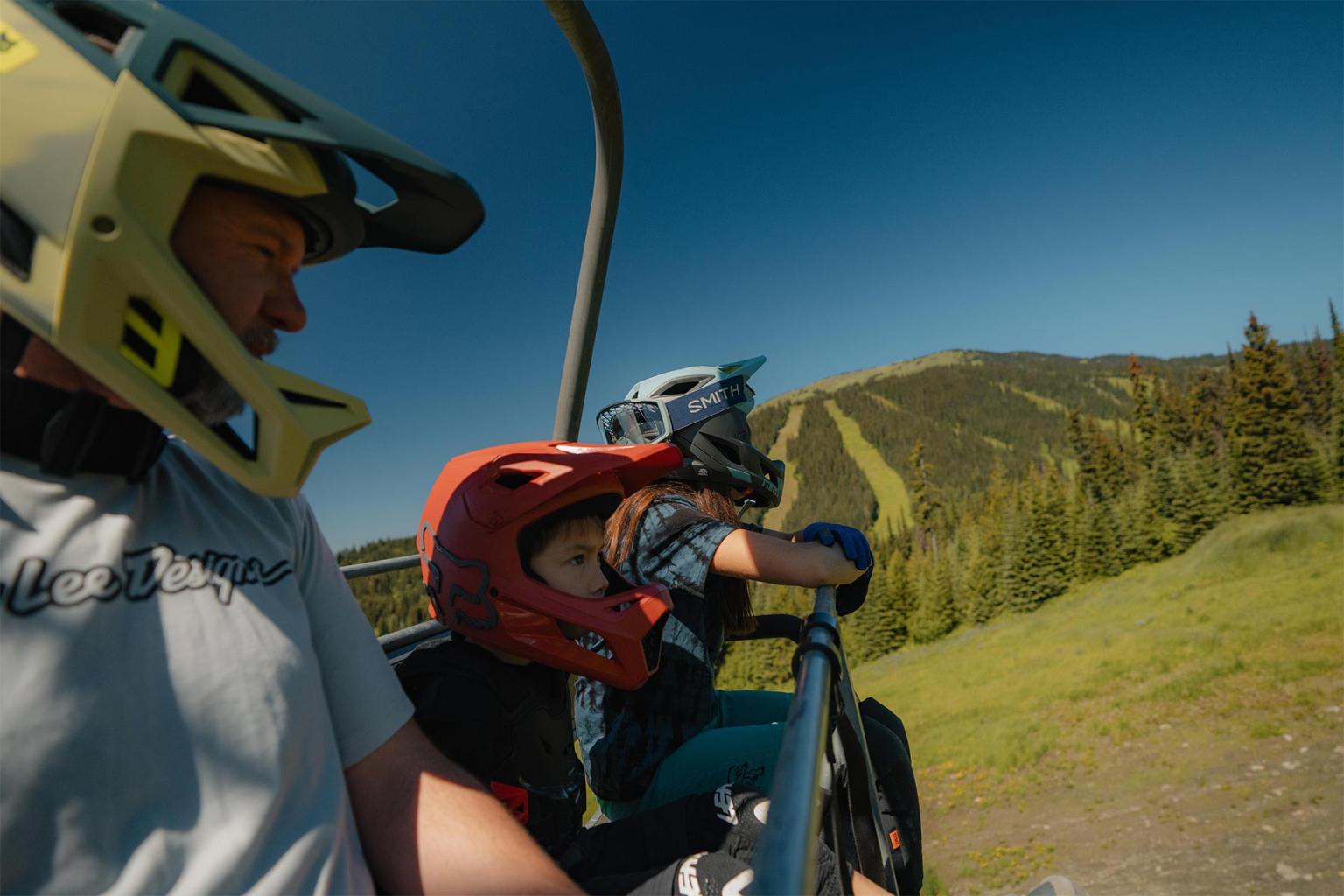 Family on a ski lift with helmets, overlooking a mountain under clear blue sky.