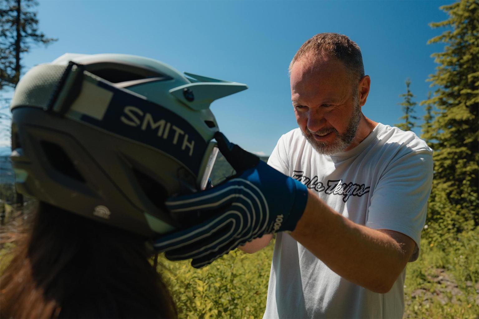 Man adjusting a helmet on a child outdoors, with trees in the background.