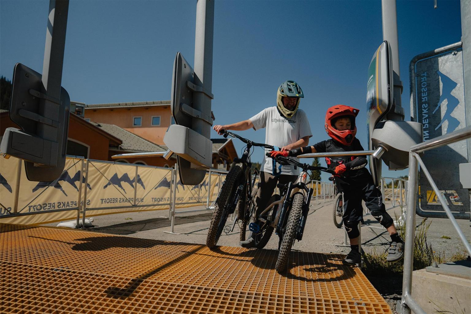 Two cyclists in helmets prepare to pass a gate on a sunny day.