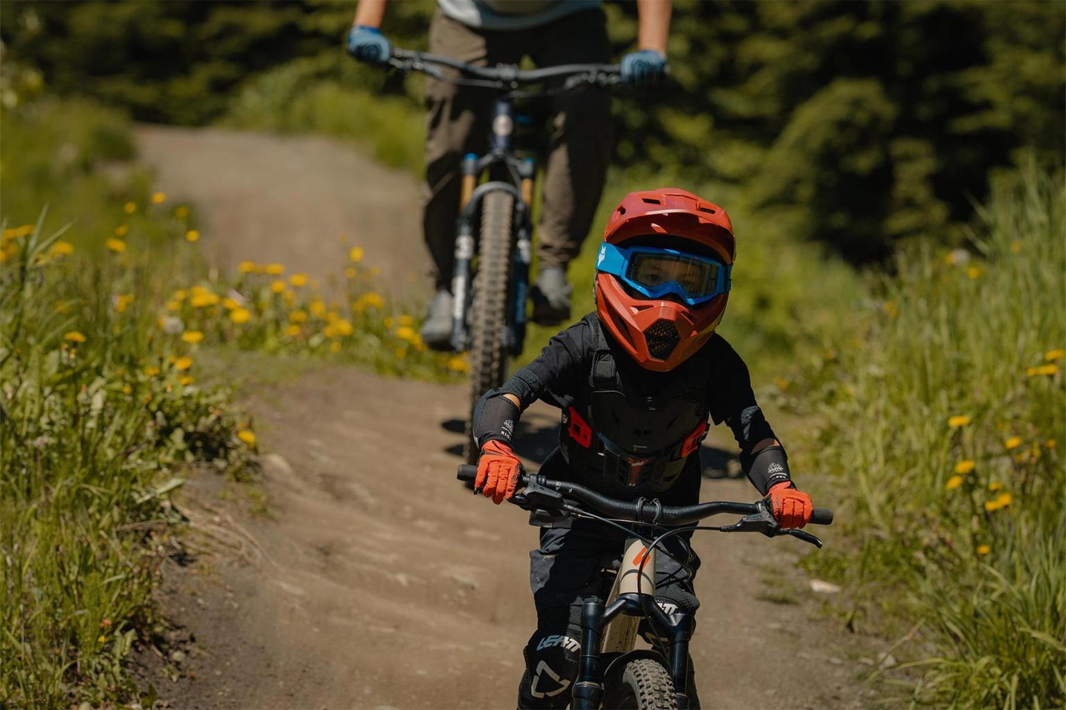 Child biking on a dirt trail, followed by an adult, both in protective gear.