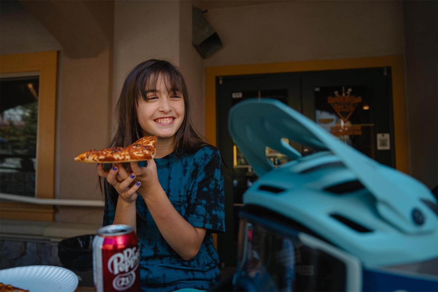 Smiling woman holds pizza slice, sitting outdoors with soda can and helmet nearby.