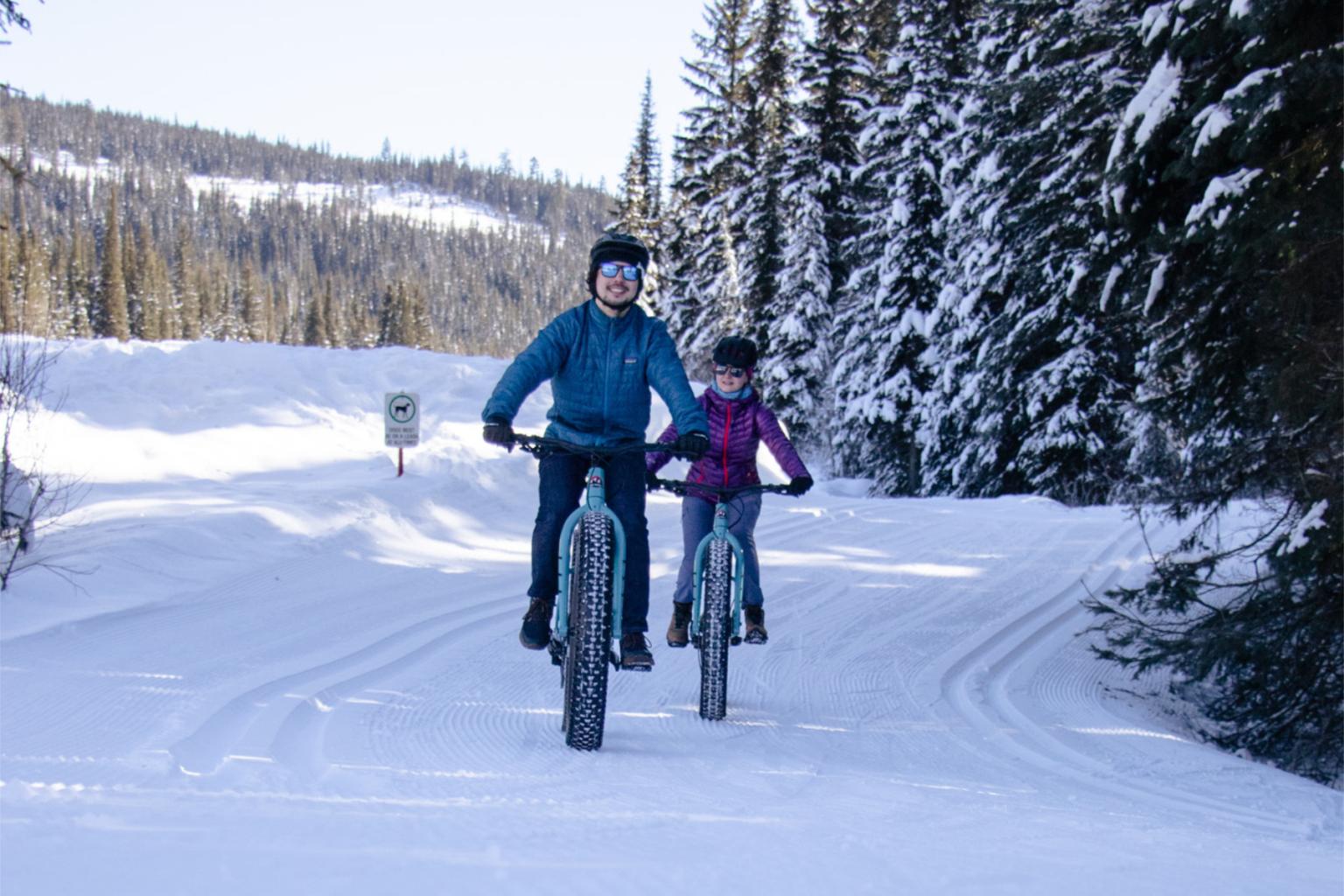 Two people ride fat bikes on a snowy forest trail.