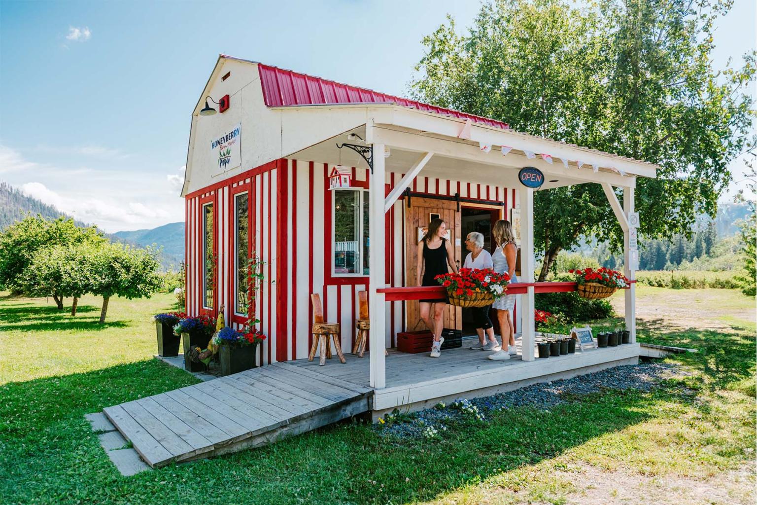 Small red and white farm stand with people on a porch, set in a sunny rural landscape.