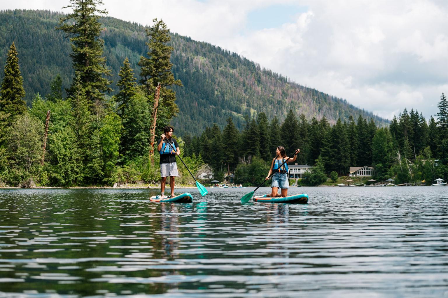 Two people paddleboarding on a lake, surrounded by trees and mountains.