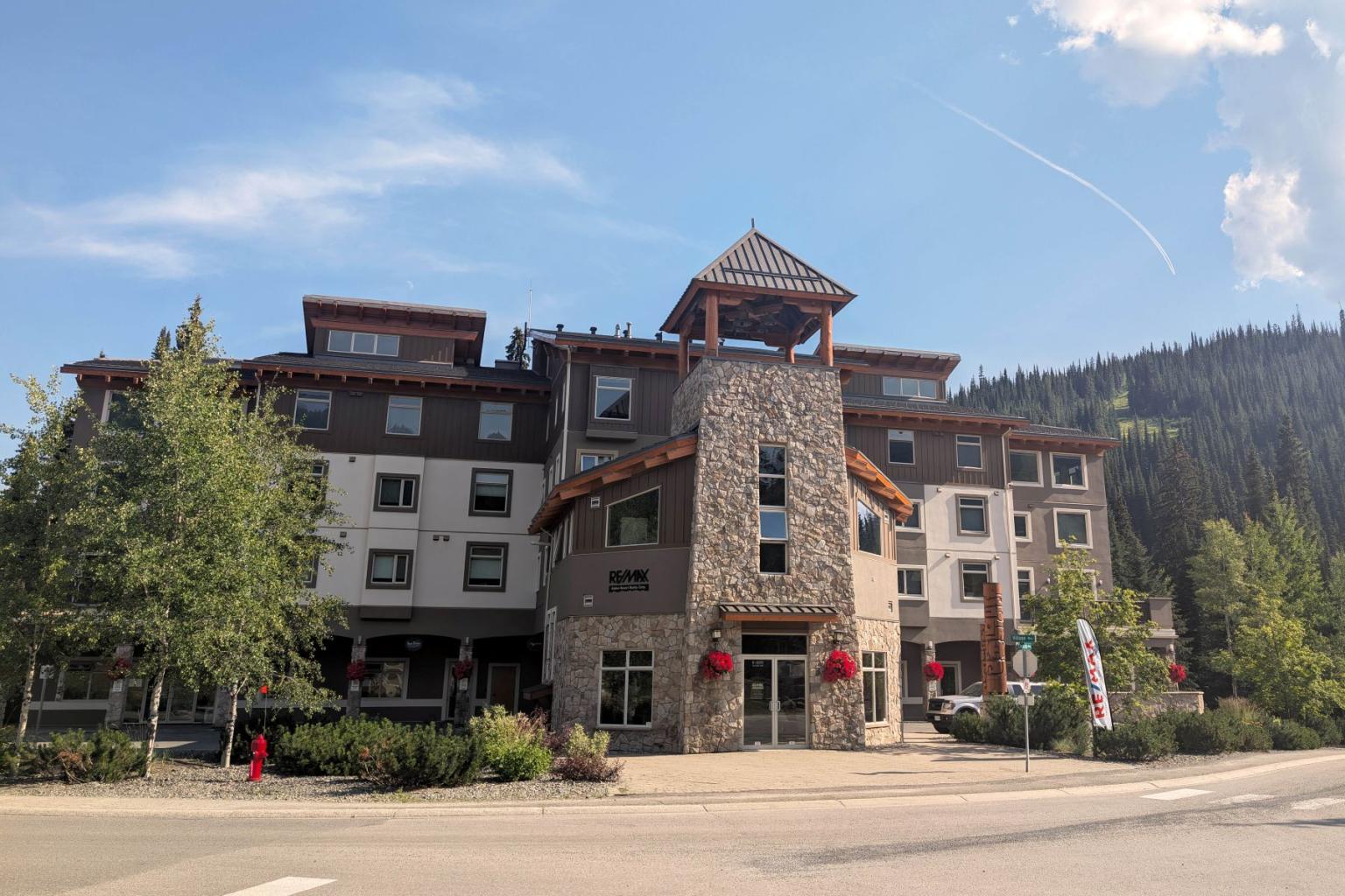 Hotel with stone facade and red flowers under a blue sky, surrounded by trees.
