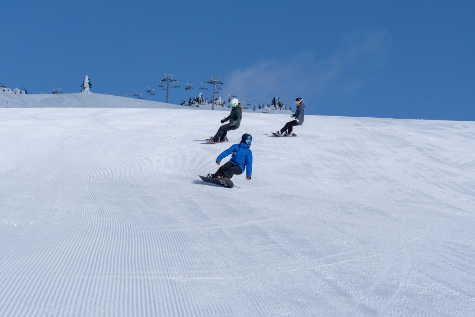 Three snowboarders carving down a snowy slope under a clear blue sky.