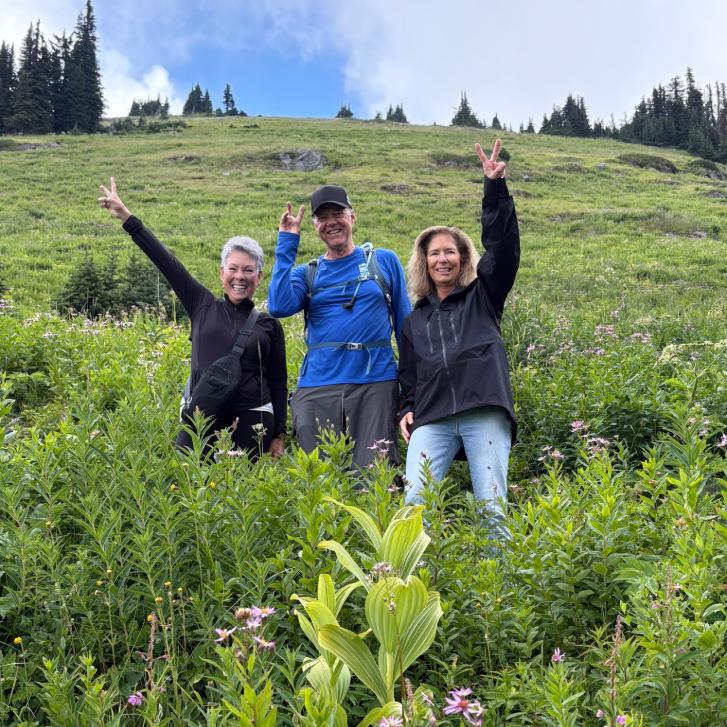 Three people smiling, posing with peace signs in a lush green alpine meadow on a hike.