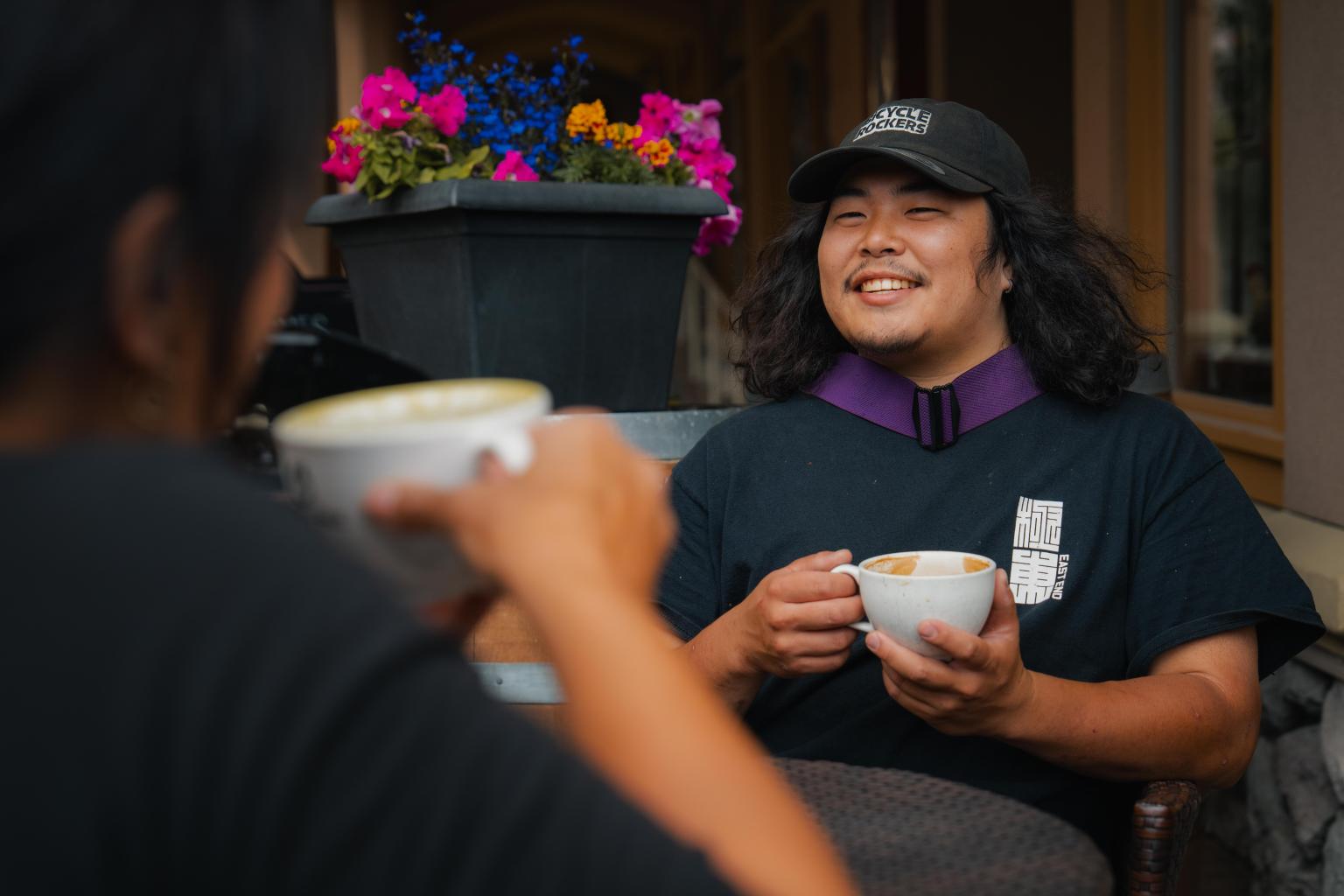 Two people smiling and drinking coffee outside, flowers in the background.