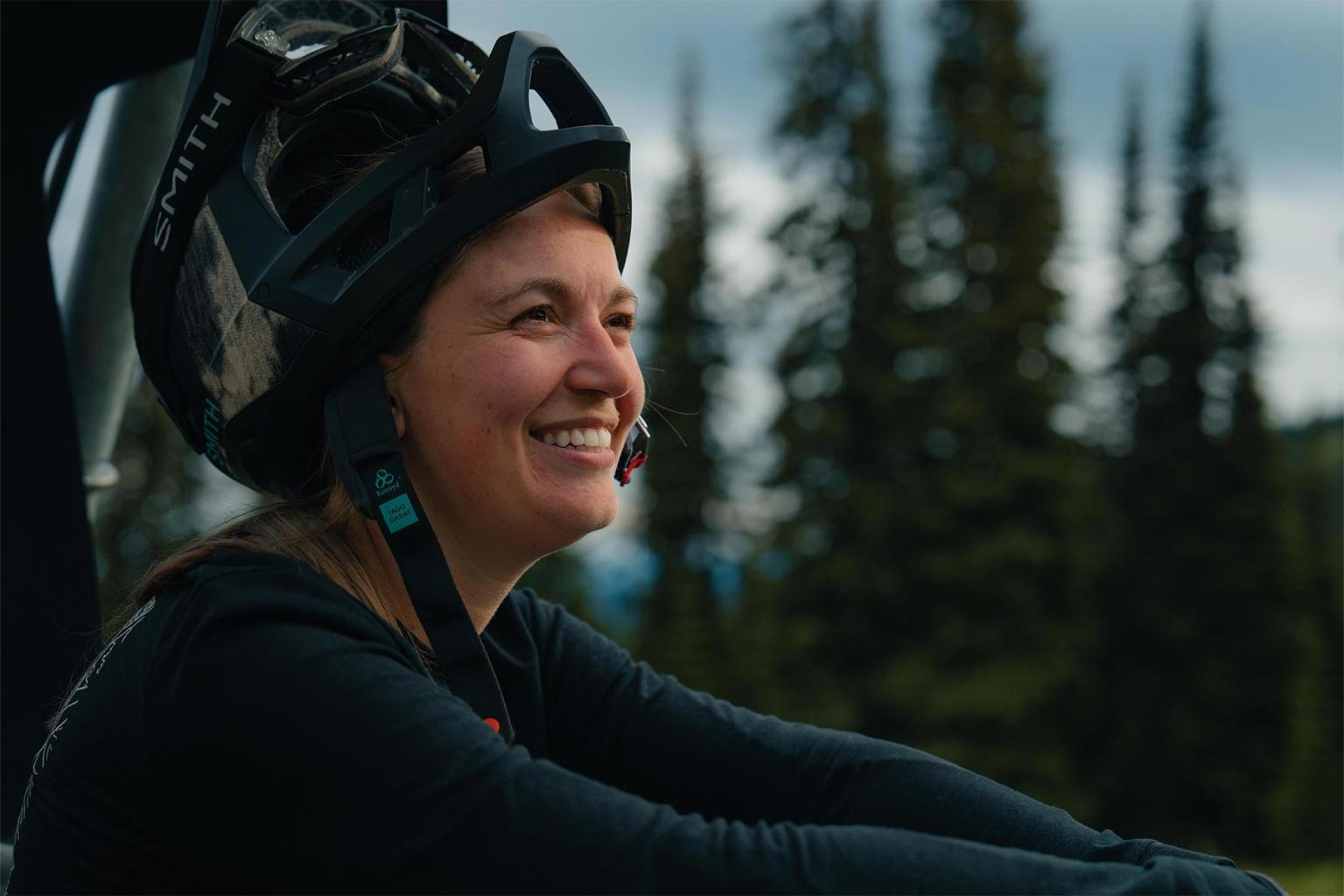 Smiling person wearing a bike helmet, sitting outdoors with trees in the background.