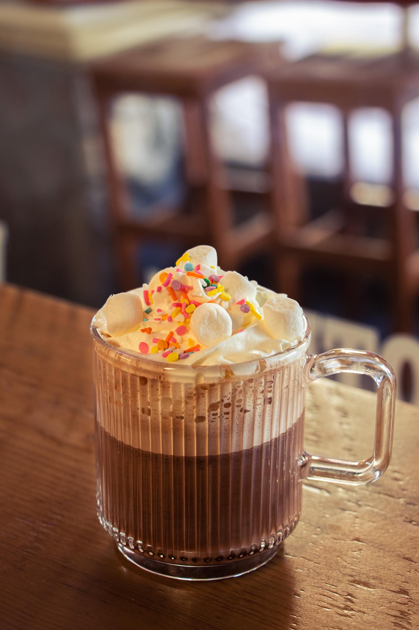 Hot chocolate topped with whipped cream, marshmallows, and colorful sprinkles in a glass mug.