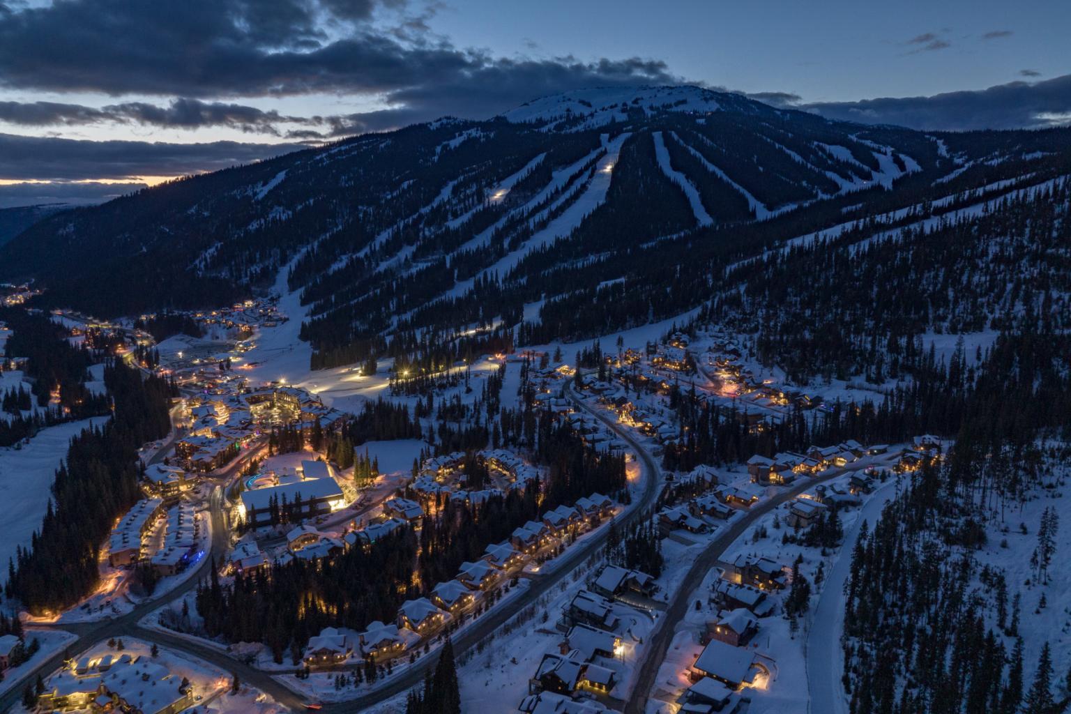 Snowy village at dusk with glowing lights, beneath a mountain with ski slopes.