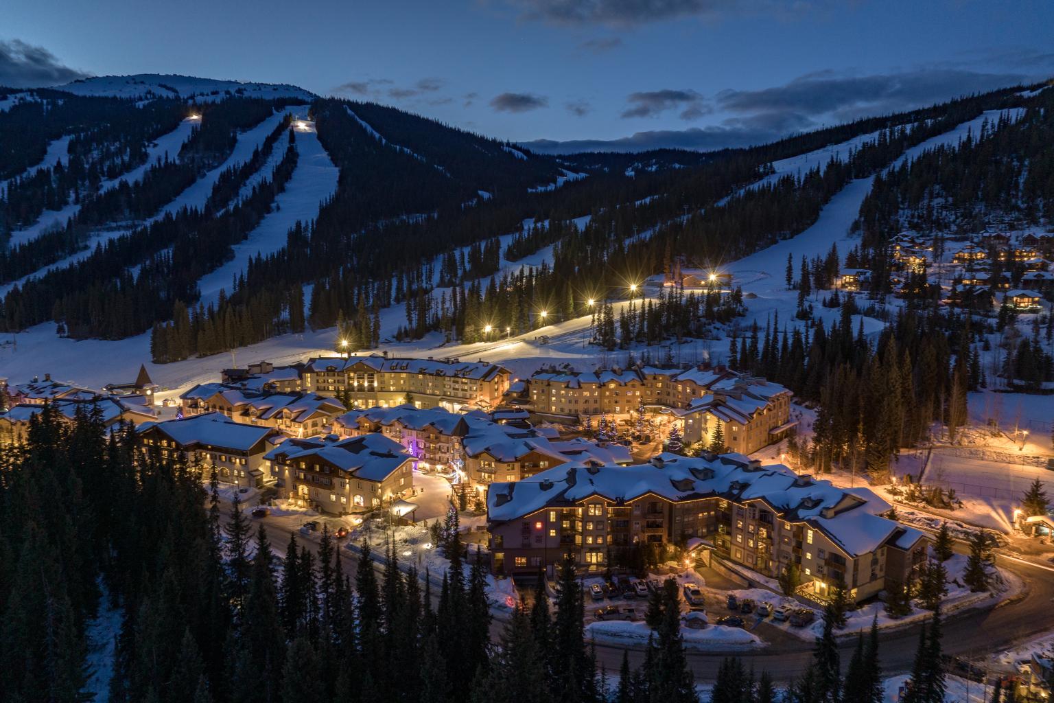 Snowy village at dusk, warm lights glowing, surrounded by ski slopes and mountains.
