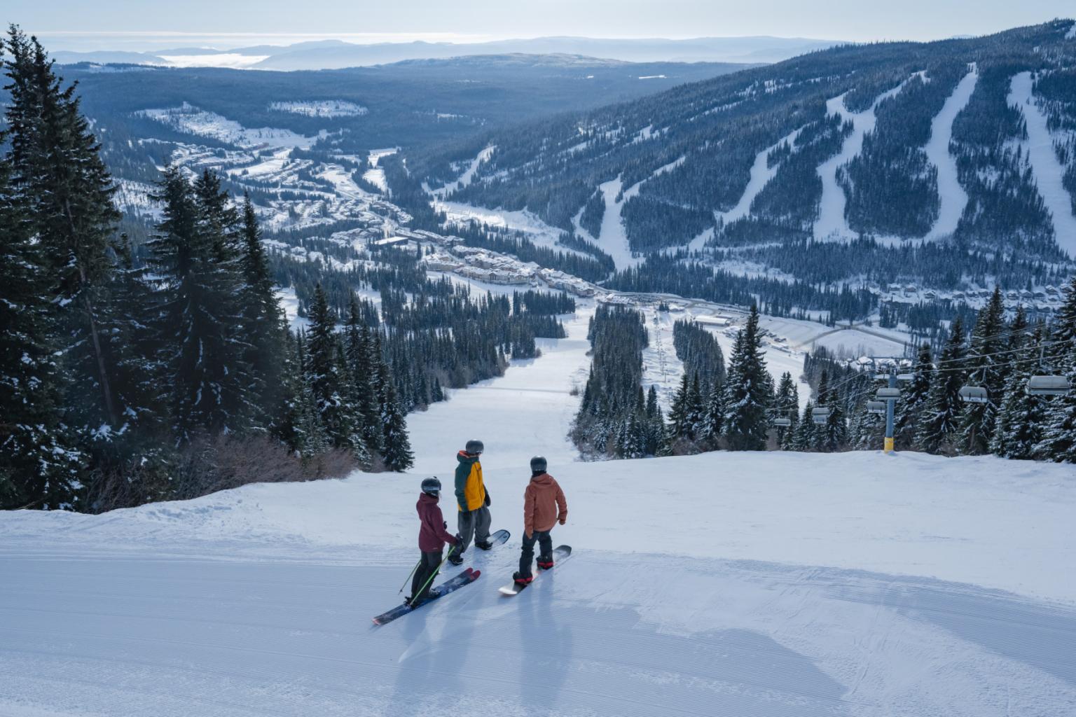 Skiers on a snowy slope, overlooking a wide valley with distant mountains.