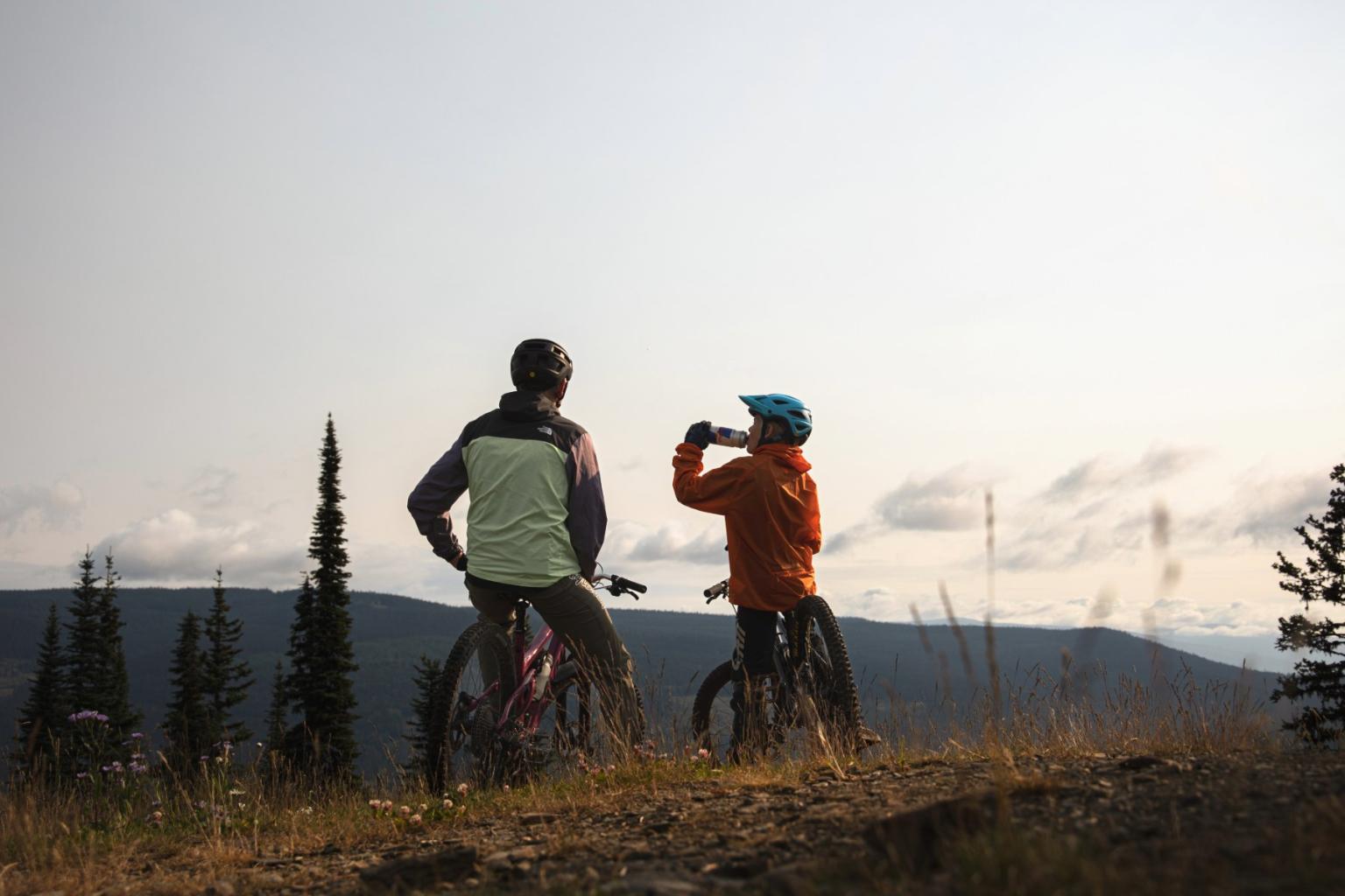 Two cyclists rest on a hilltop, one drinking water, overlooking mountains.