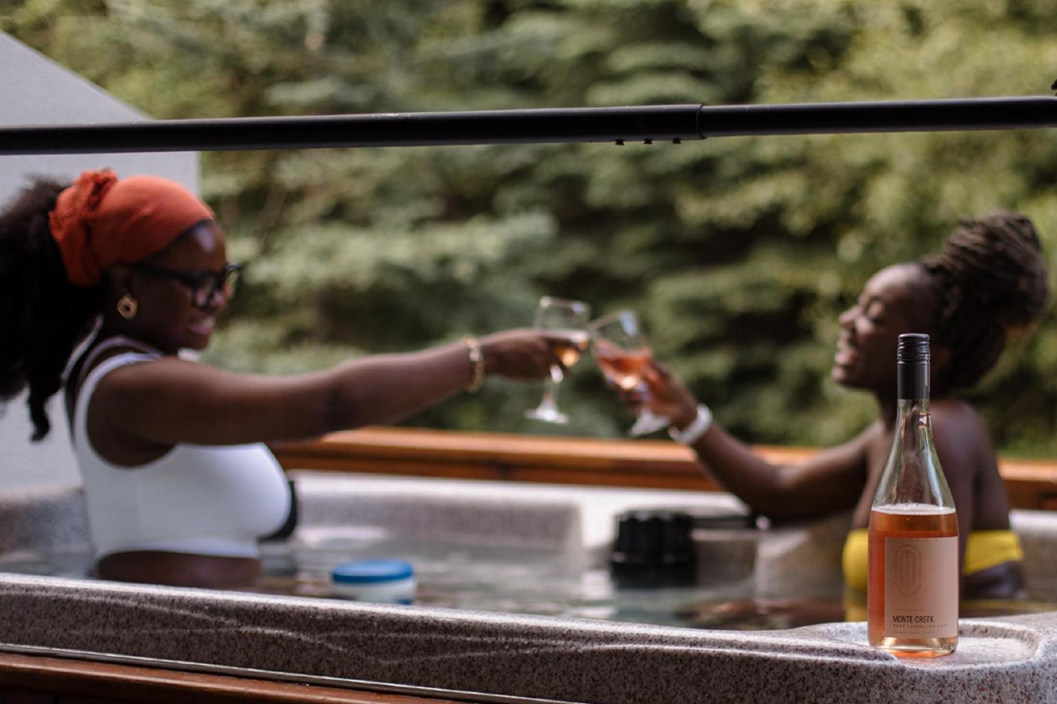 Two people clinking wine glasses in a hot tub, surrounded by trees.
